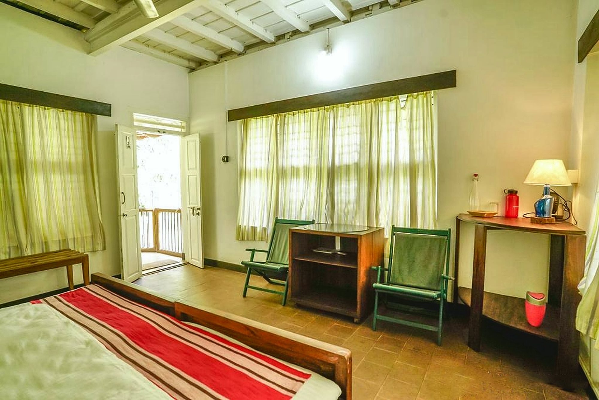 A bedroom with white double doors open onto a wooden balcony and bright greenery